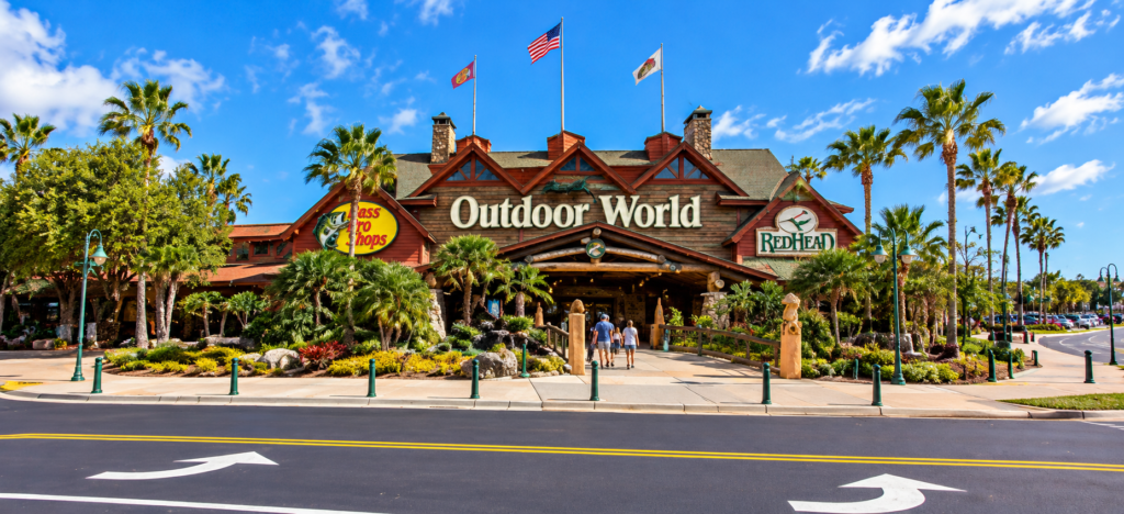 Imagem ilustrativa da Bass Pro Shops Outdoor World em Orlando, com fachada rústica de madeira, palmeiras e entrada principal da loja.