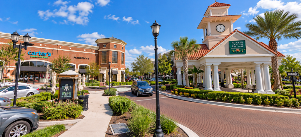 Imagem ilustrativa do Winter Garden Village, próximo a Orlando, com torre do relógio, lojas, palmeiras e ruas arborizadas.